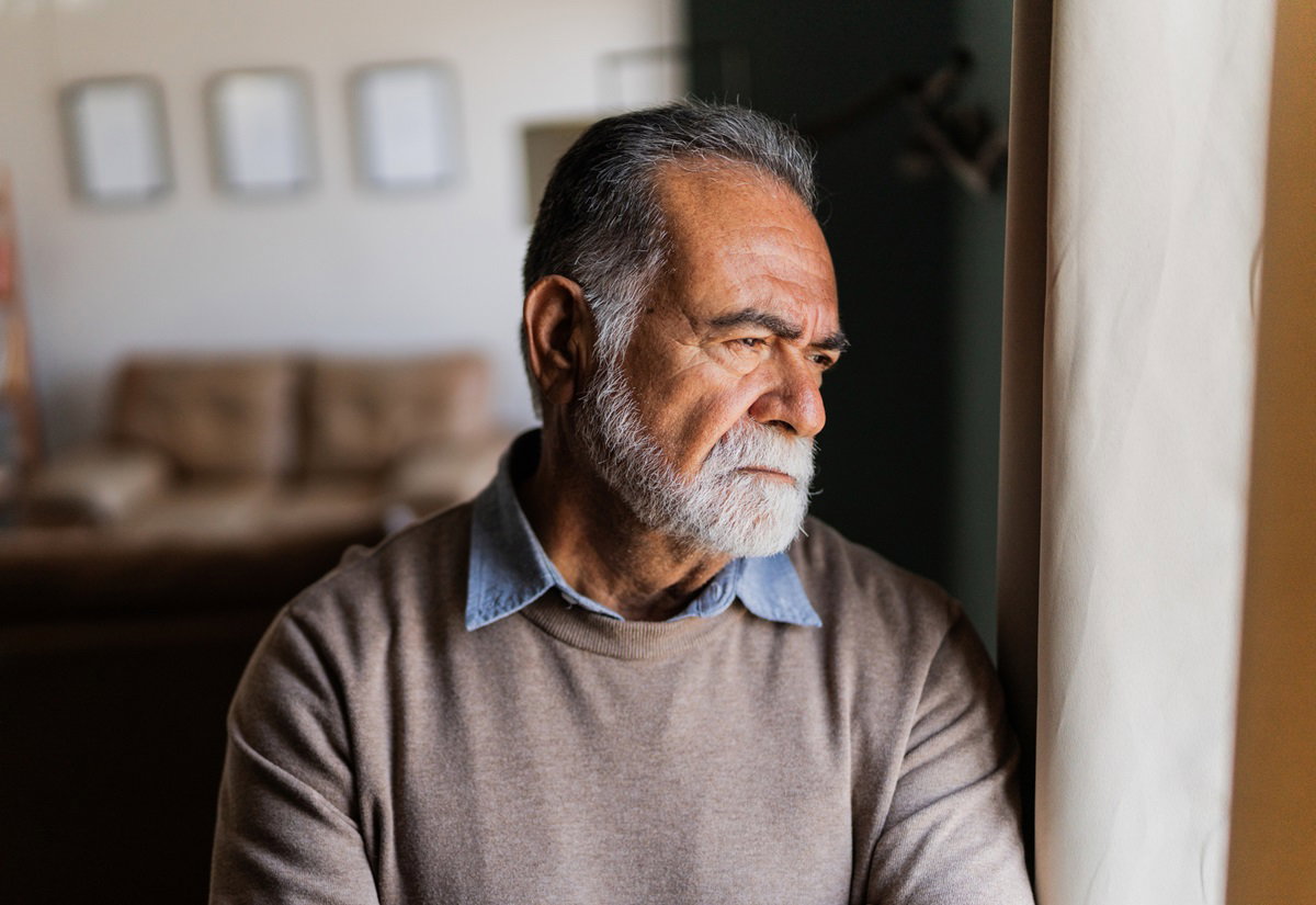 starszy mężczyzna senior z siwą brodą i wąsami ubrany w koszulę i sweter patrzy smutno w okno