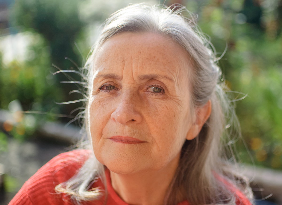 kobieta seniorka z dłuższymi siwymi włosami i w czerwonym swetrze jest zewnątrz i patrzy zamyślona