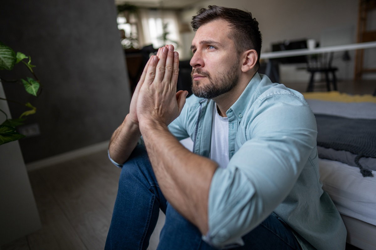 młody mężczyzna siedzi na podłodze i ma dłonie złożone do modlitwy patrząc w dal zrozpaczonym wzrokiem