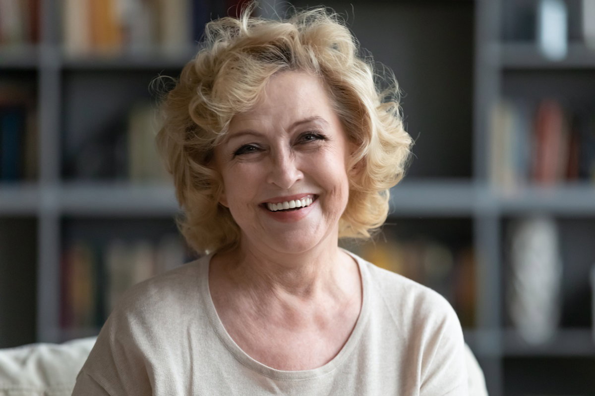 kobieta w dojrzałym wieku seniorka z blond lokami siedzi w salonie i uśmiecha się szeroko