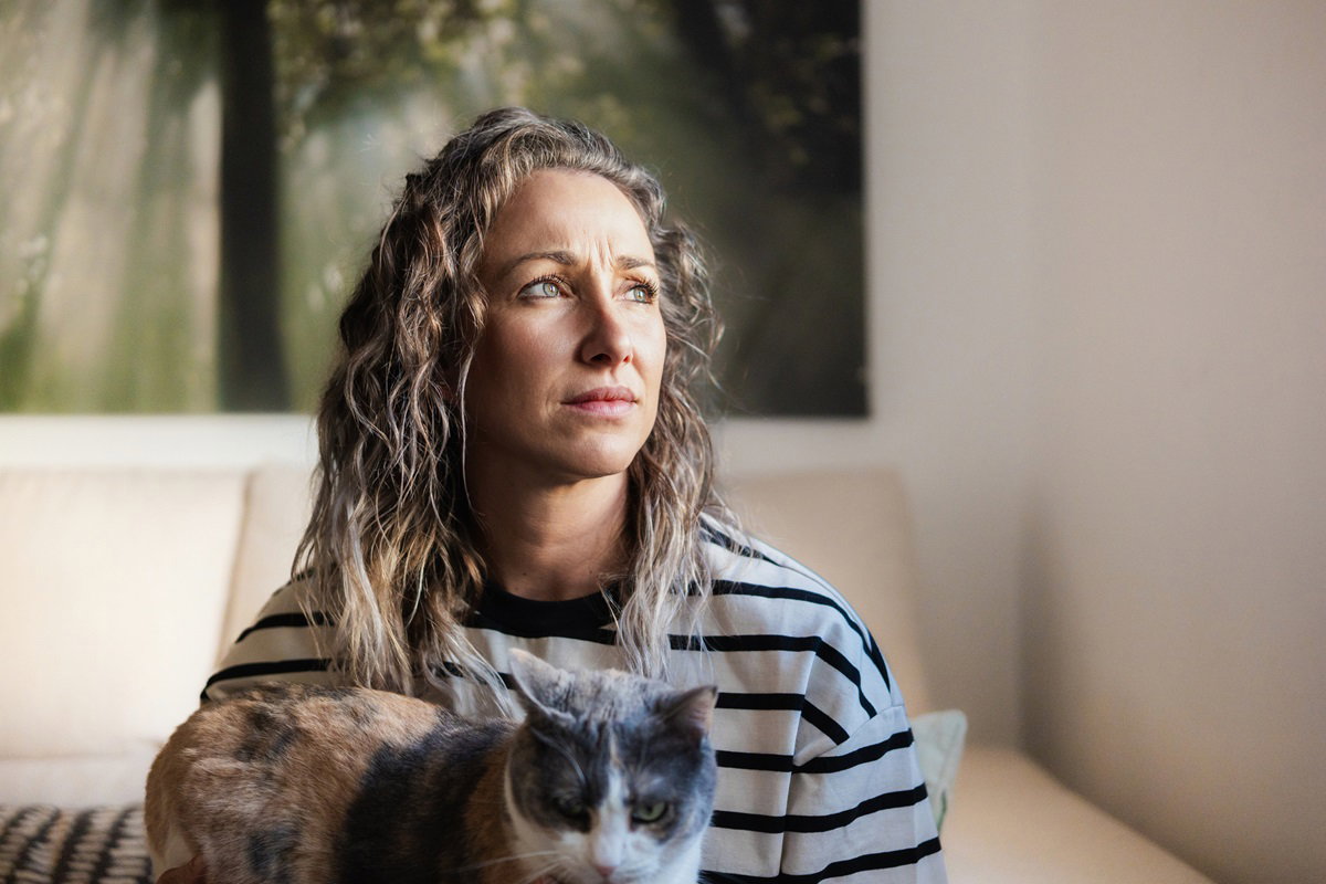 młoda kobieta z pofalowanymi włosami i kotem na kolanach siedzi i patrzy zamyślona w dal