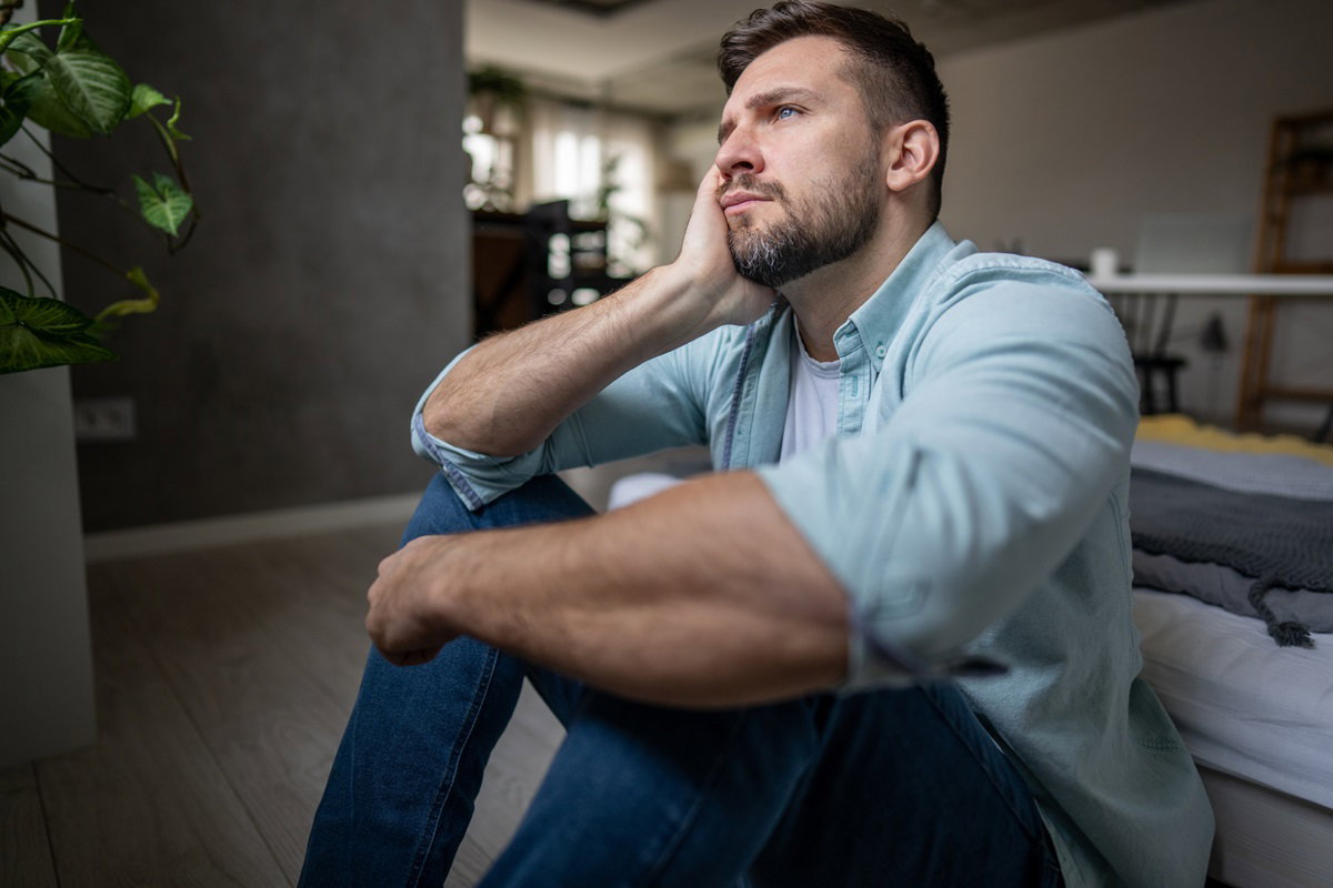 mężczyzna z brodą w średnim wieku siedzi na podłodze i ręką podpiera głowę patrząc ze smutkiem przed siebie