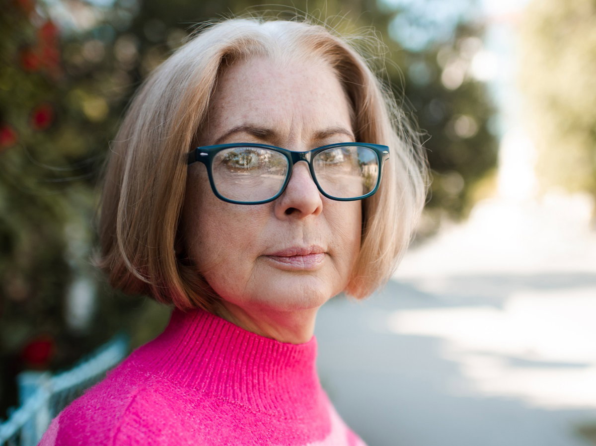 kobieta w senioralnym wieku koło 50tki w okularach i różowym swetrze stoi w ogrodzie i patrzy poważnym wzrokiem