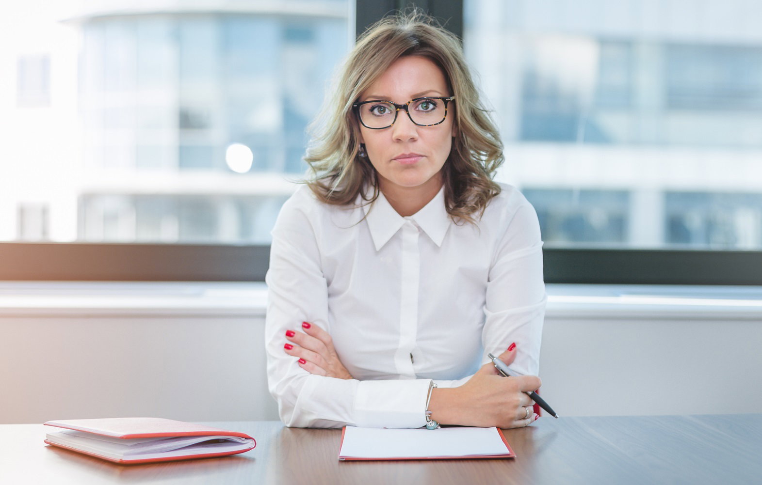 kobieta w wieku około 40 lat w białej koszuli i okularach siedzi w biurze przy biurku z założonymi rękami i ze zmartwioną miną