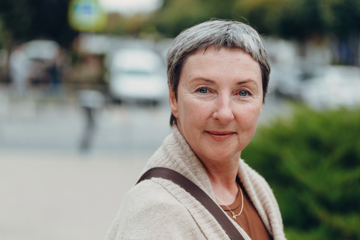kobieta w senioralnym wieku 60+ krótko ostrzyżona siwa fryzurka stoi na zewnątrz i uśmiecha się uroczo