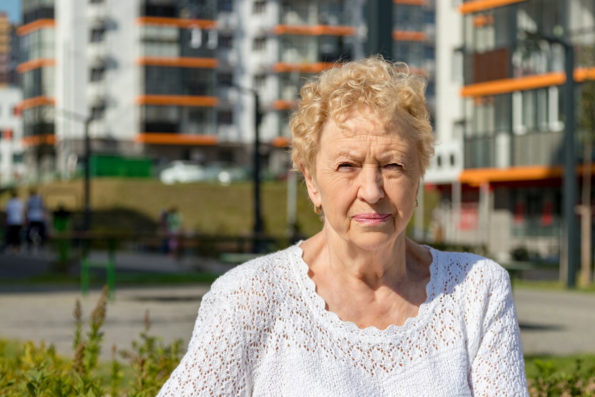kobieta w wieku 60+ w ażurowym białym sweterku i w blond loczkach stoi z zaciętą miną na tle osiedla