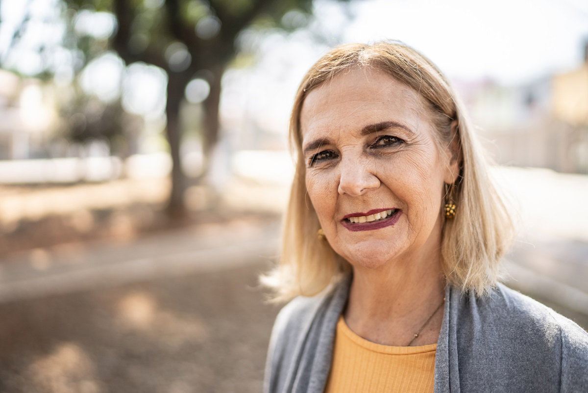kobieta w wieku 60+ seniorka z blond włosami i w makijażu oraz kolczykach stoi na zewnątrz i uśmiecha się niepewnie