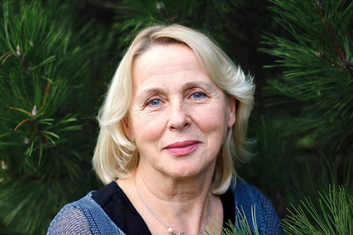 kobieta w dojrzałym wieku z blond włosami stoi na tle sosen i lekko się uśmiecha