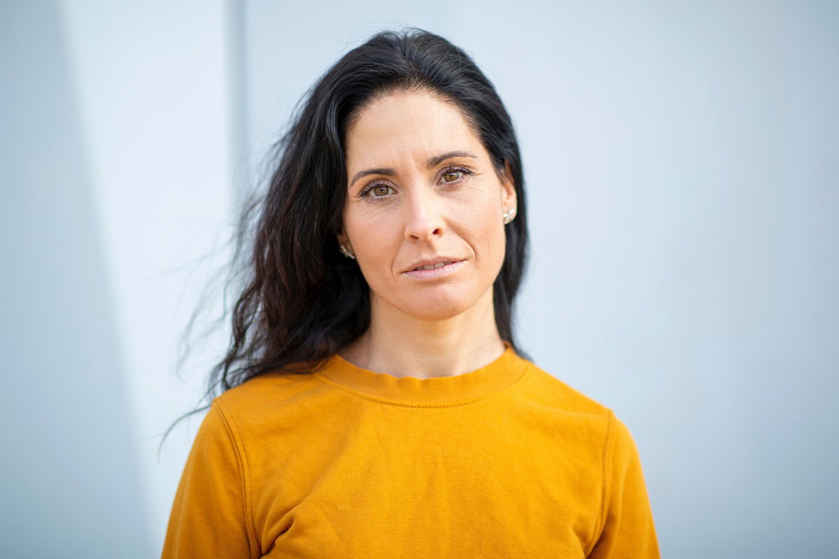 kobieta w średnim wieku z ciemnymi włosami i w żółtej bluzce stoi i patrzy przed siebie poważnie