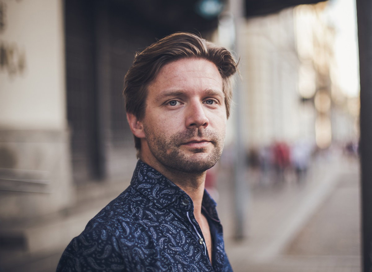 młody mężczyzna blondyn stoi na ulicy w koszuli we wzorki i patrzy poważnym wzrokiem