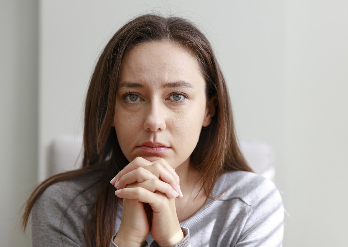 młoda dziewczyna lat około 30 z długimi włosami siedzi smutna w fotelu na tle ściany ze splecionymi rękami