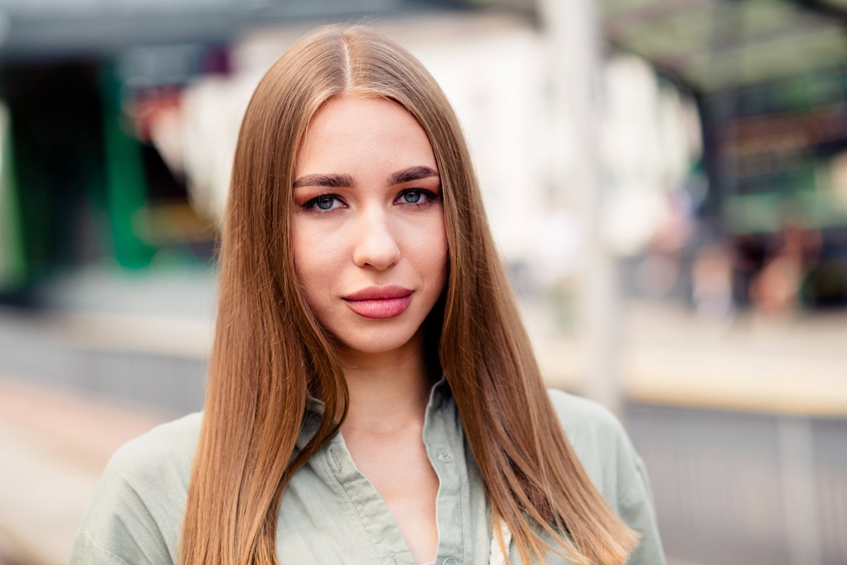 młoda dziewczyna w jasnozielonej bluzce z jasnymi długimi włosami i w makijażu stoi na ulicy z poważną miną i patrzy przed siebie