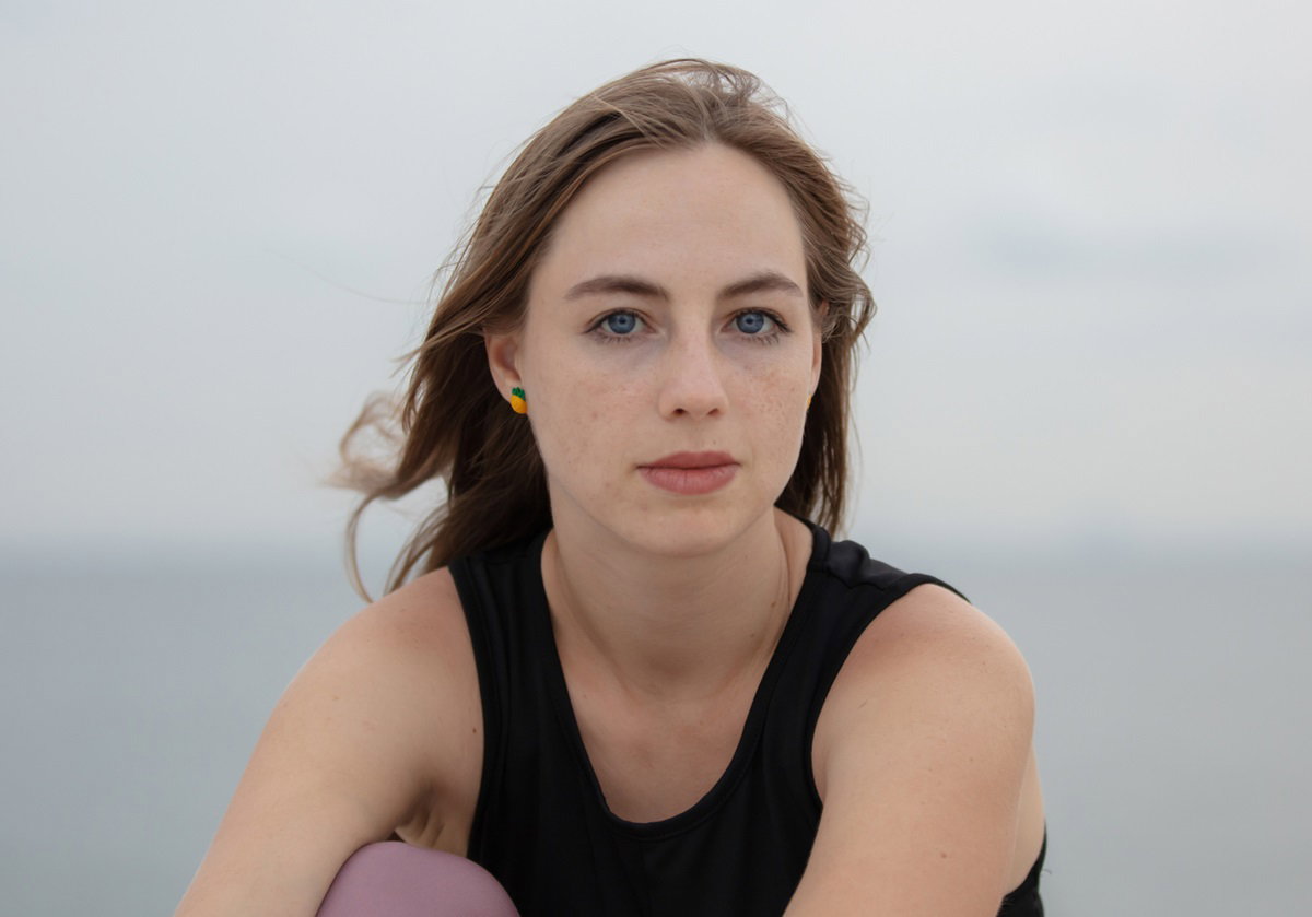 młoda dziewczyna z długimi włosami w czarnym topie siedzi na plaży na tle morza i patrzy smutnym wzrokiem