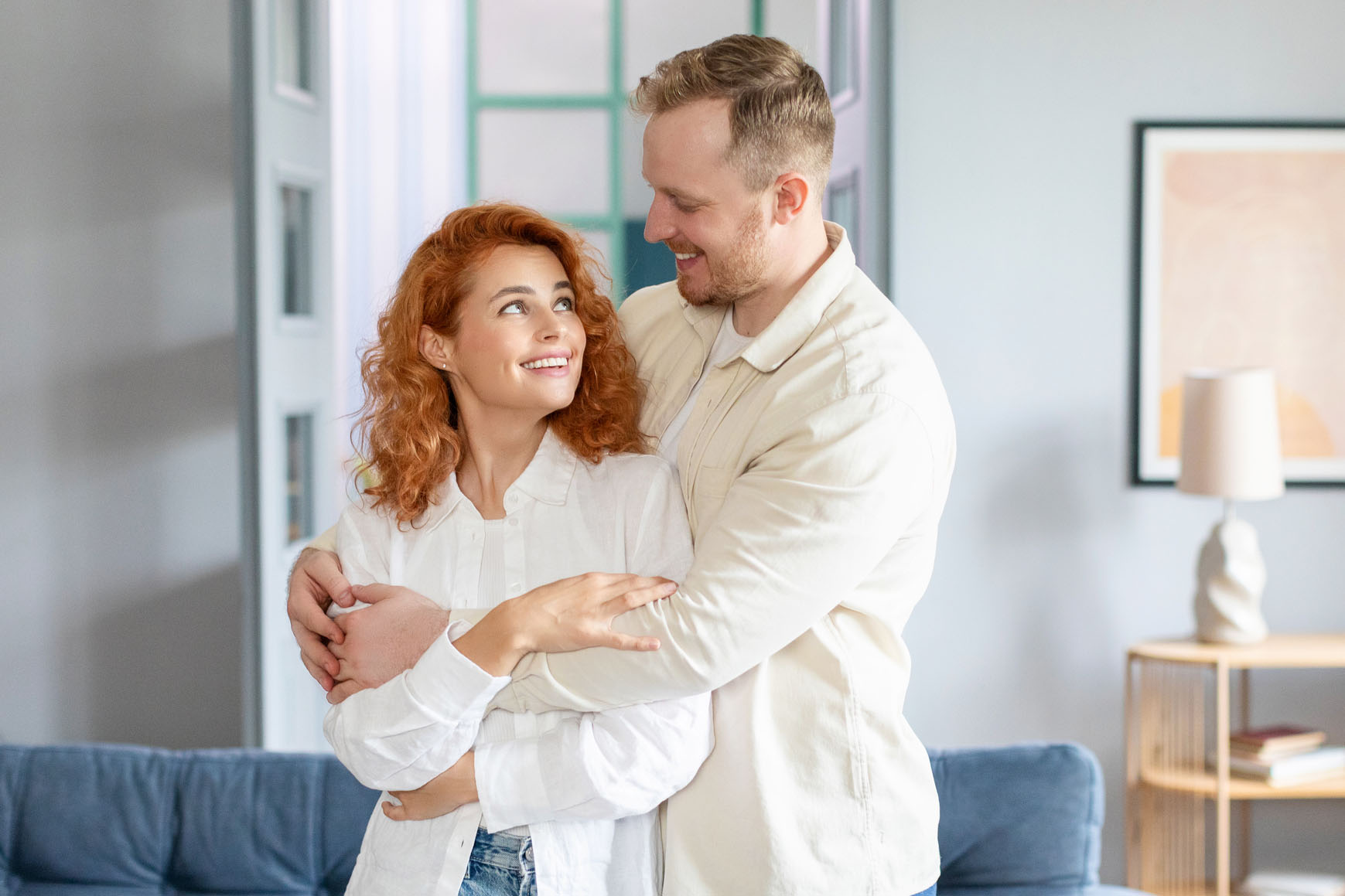 Loving European couple cuddling while standing in modern living room, enjoying relocation, relish talk and harmonic relationships. New apartment owners on moving day
