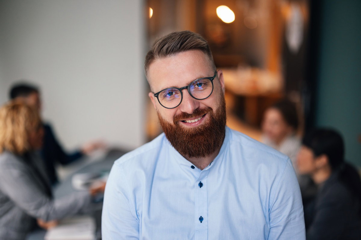 mężczyzna w wieku około 40 lat w jasnej koszuli i z rudą brodą i w okularach stoi w pracy i uśmiecha się lekko