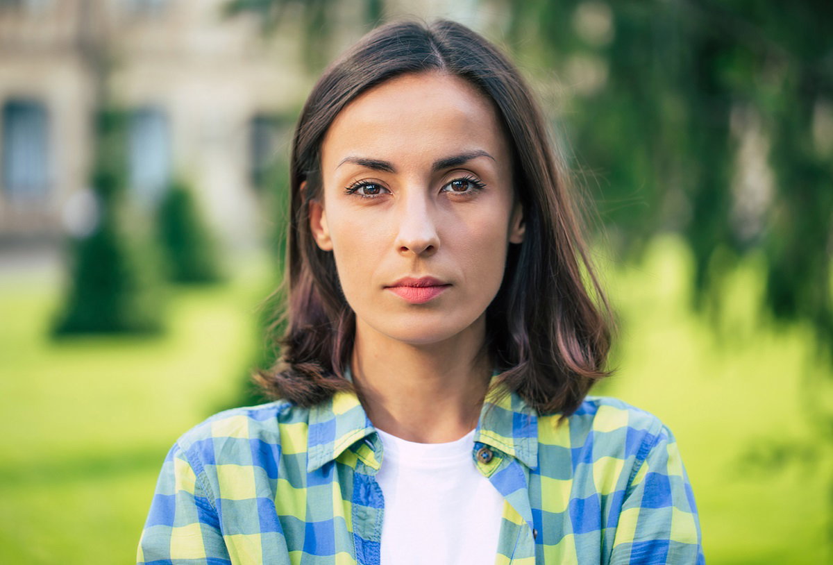 młoda kobieta w koszuli w niebiesko-żółtą kratkę stoi na zewnątrz i patrzy poważnym wzrokiem przed siebie
