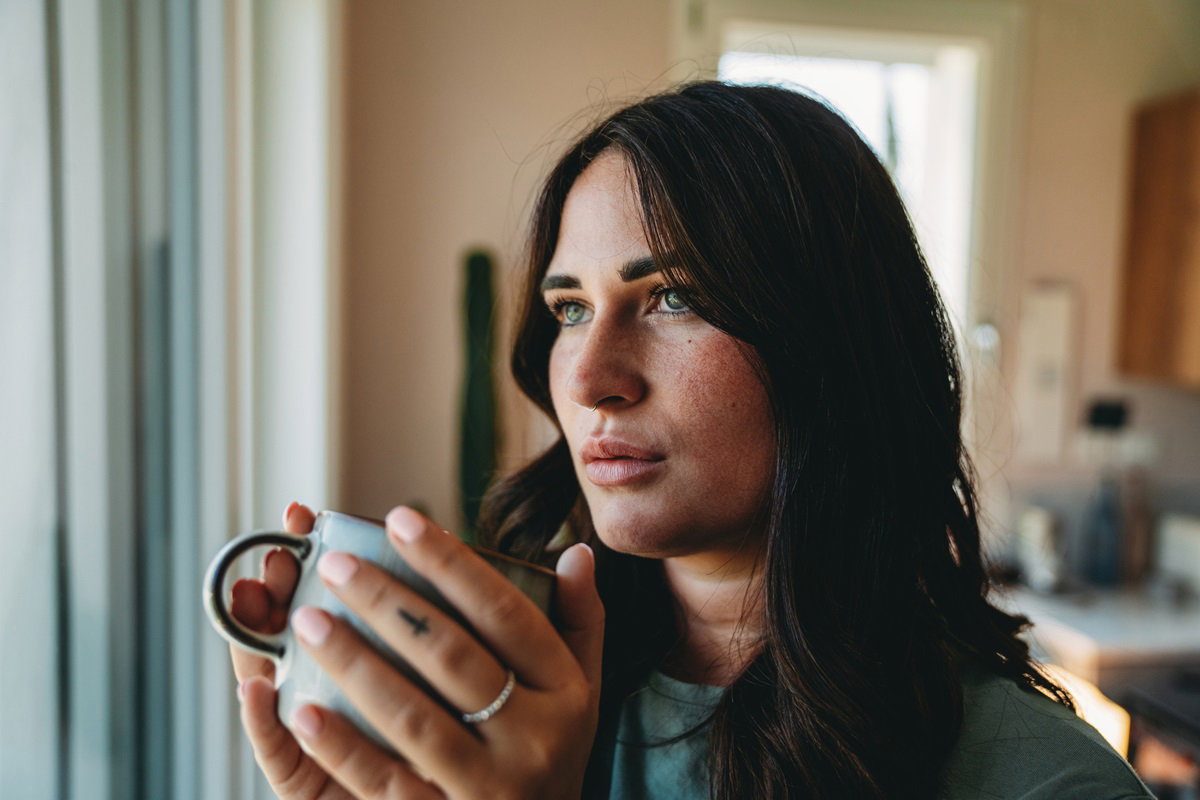 młoda dziewczyna z ciemnymi długimi włosami patrzy w okno zamyślona trzymając w rękach kubek