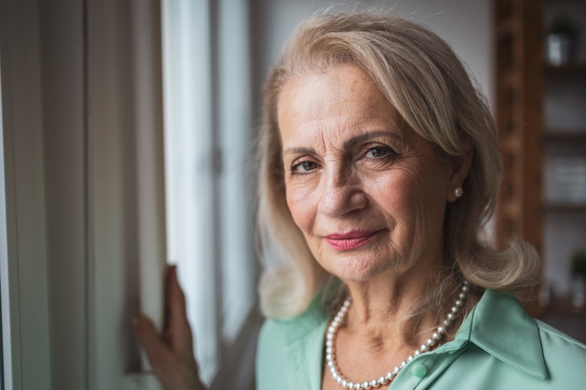kobieta w senioralnym wielu 60+ blondynka z perłami i w zielonej bluzce stoi przy framudze i patrzy gorzkim spojrzeniem
