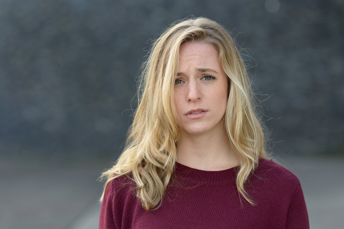 młoda dziewczyna z długimi blond włosami i w bordowym sweterku stoi na zewnątrz ze zmartwioną miną