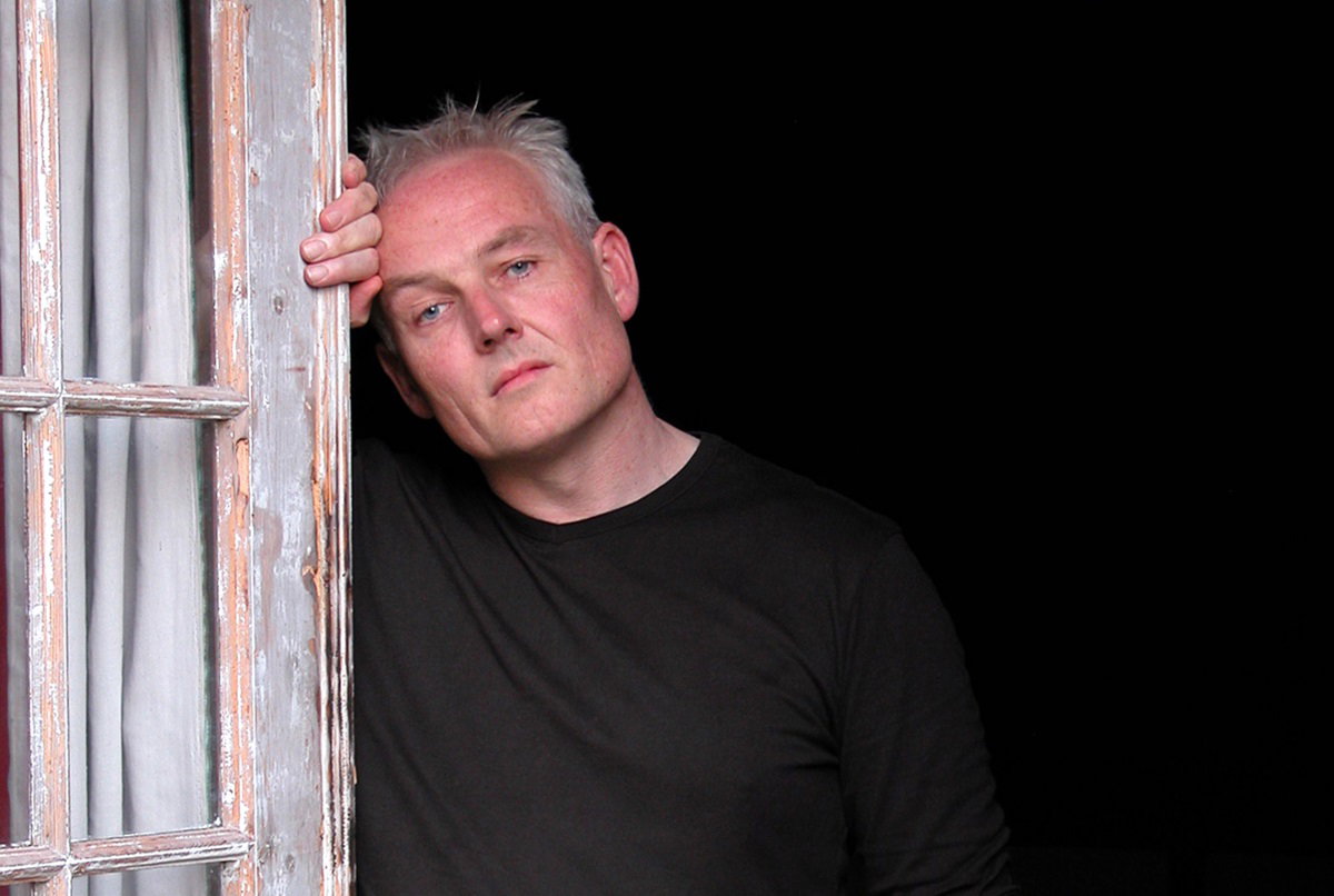 mężczyzna w średnim wieku z siwymi włosami opiera się o drewniane drzwi i patrzy zamyślonym wzrokiem