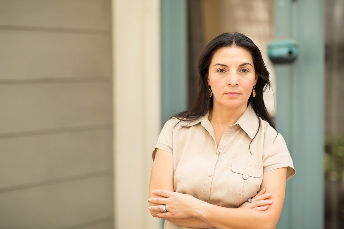 kobieta w średnim wieku około 40 lat z czarnymi długimi włosami stoi na tle domu z założonymi rękami i zaciętą miną