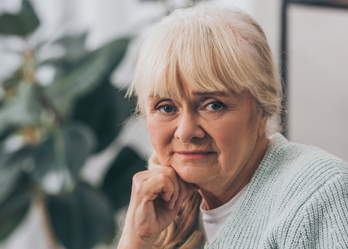 kobieta seniorka z blond włosami i grzywka w jasnym swetrze siedzi w salonie na tle kwiatka i ma zmartwioną minę