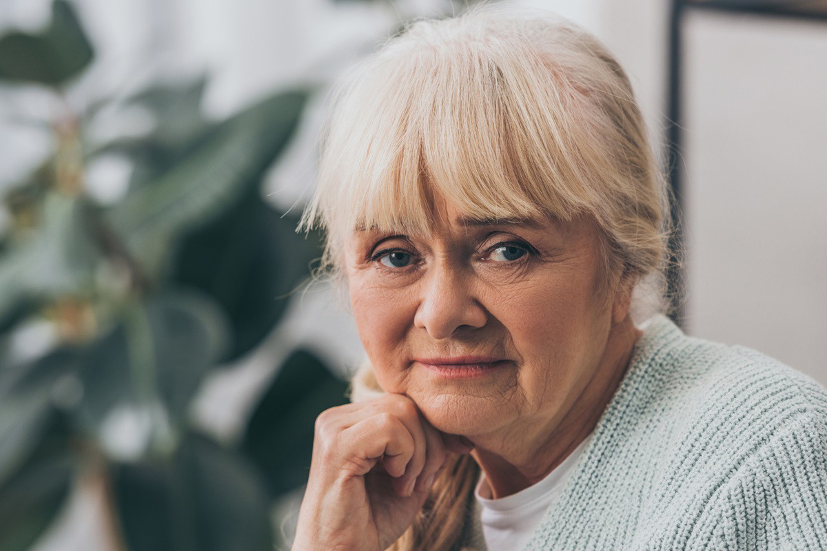 kobieta seniorka z blond włosami i grzywka w jasnym swetrze siedzi w salonie na tle kwiatka i ma zmartwioną minę