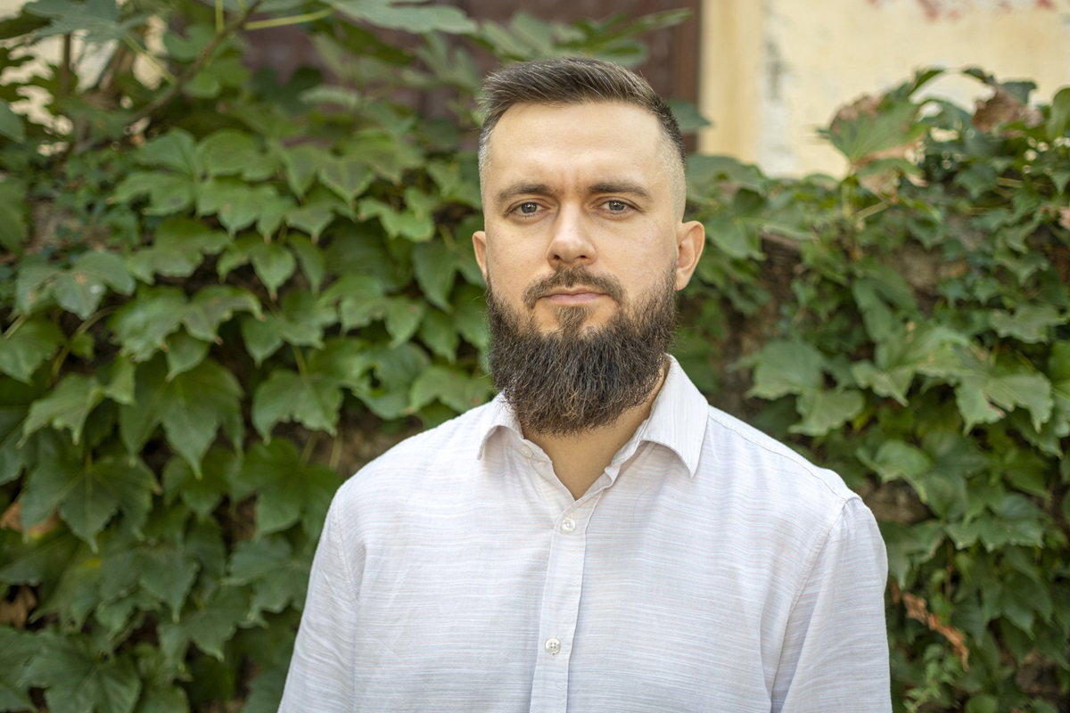 mężczyzna w wieku około 40 lat z długą brodą i w białej koszuli stoi na tle ściany z liści ze smutną miną