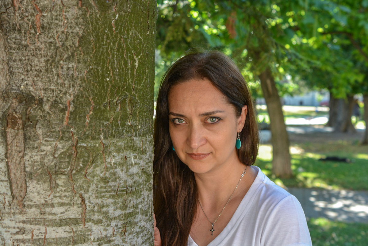kobieta w wieku około 40 lat w białym t-shircie z długimi włosami i turkusowymi kolczykami opiera się o pień drzewa i patrzy smutnym wzrokiem