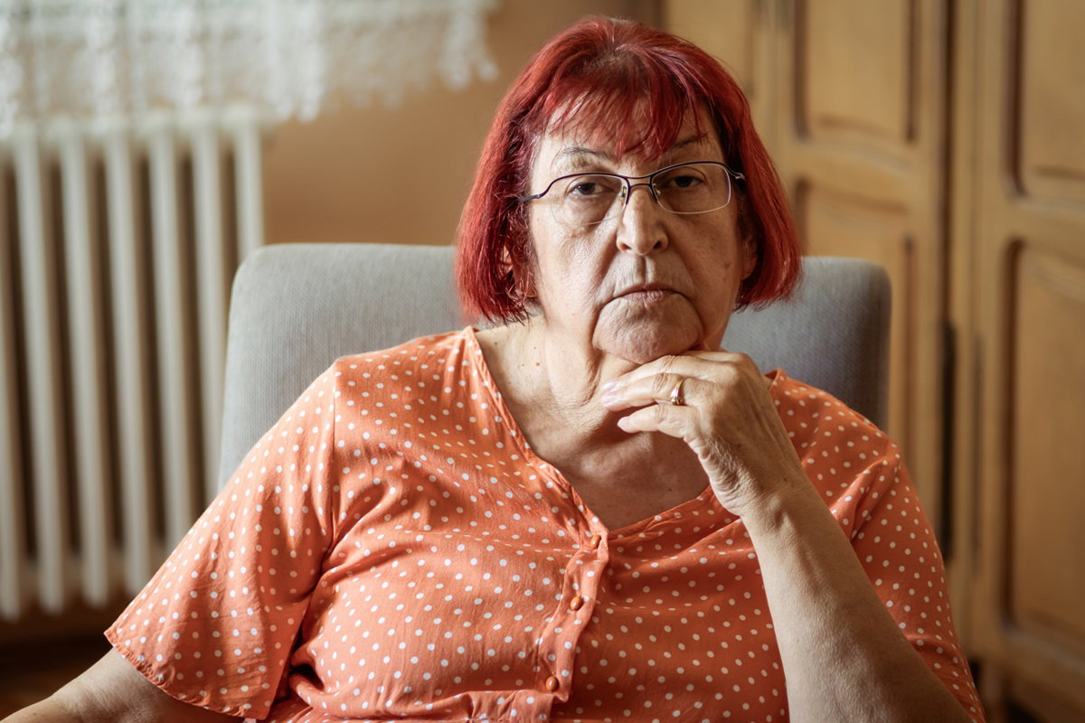 kobieta w okularach w wieku 60+ z pofarbowanymi na intensywny kolor włosami i w pomarańczowej sukience w groszki siedzi na fotelu w pokoju z poważną miną