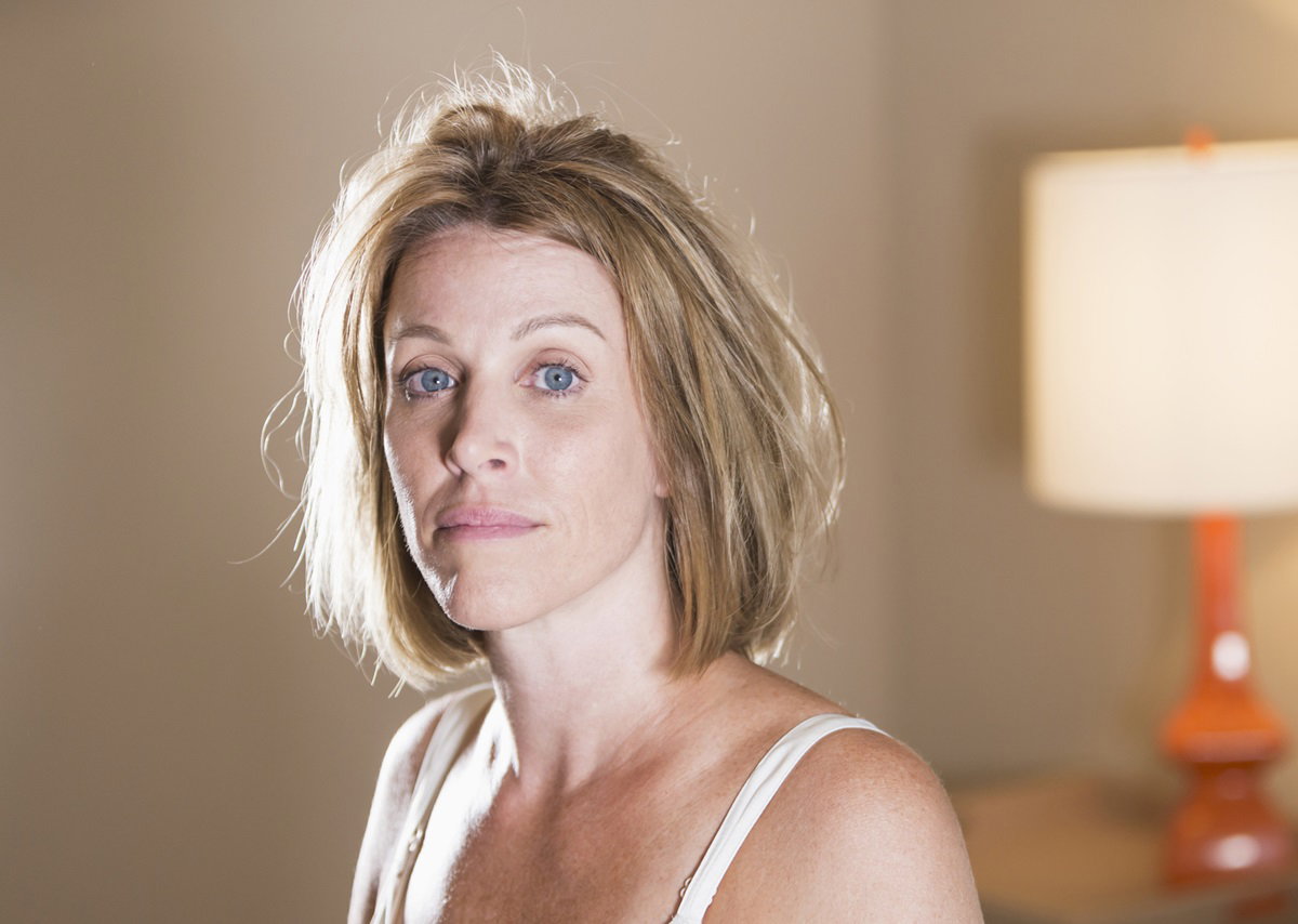 kobieta w dojrzałym wieku z jasnymi lekko rozwichrzonymi włosami siedzi na łóżku i patrzy smutno