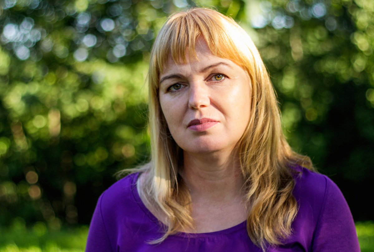 kobieta w średnim wieku około 40+ z blond włosami z grzywką i w fioletowej bluzce siedzi na ławce w parku i jest smutna