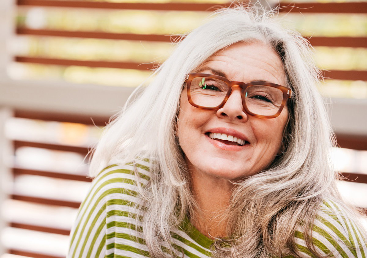 kobieta w wieku 60+ z siwymi długimi włosami i w okularach ubrana w t-shirt w zielone paski uśmiecha się z rozkoszą