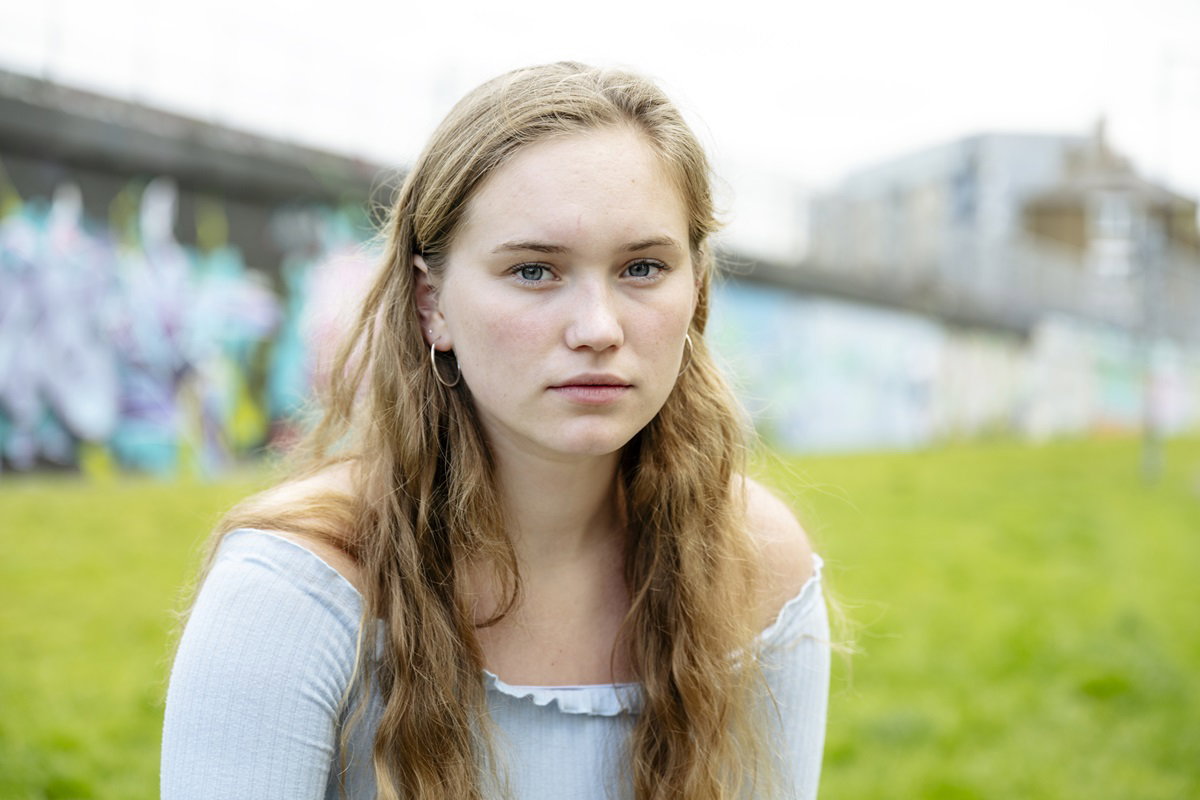 młodziutka dziewczyna z jasnymi długimi włosami siedzi na zewnątrz i patry smutno przed siebie