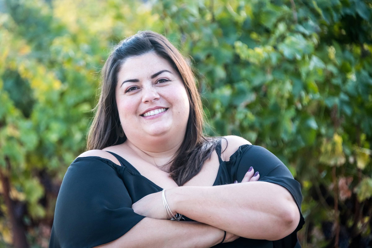 młoda kobieta w wieku około 30 lat w czarnej sukience z długimi włosami stoi na zewnątrz z założonymi rękami i się uśmiecha od ucha do ucha