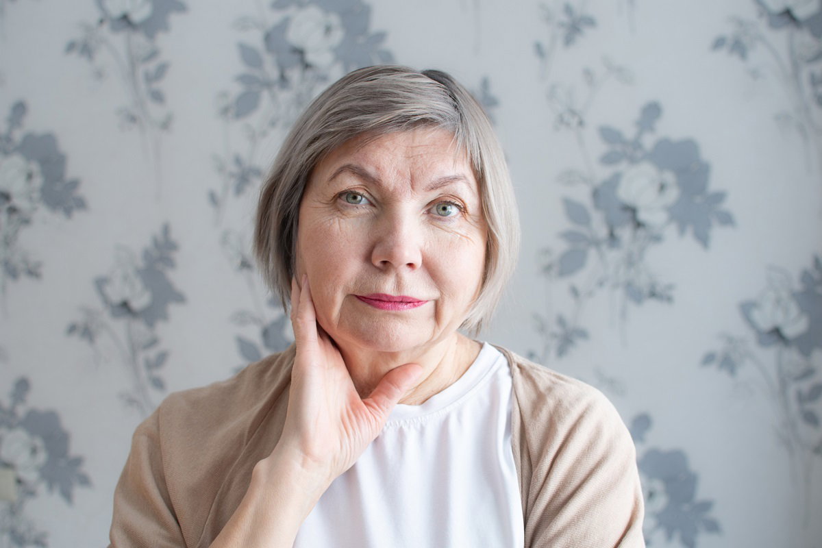kobieta w wieku 60+ z jasnymi krótko przyciętymi włosami i w beżowym sweterku siedzi na tle tapety i patrzy z powątpiewaniem przed siebie