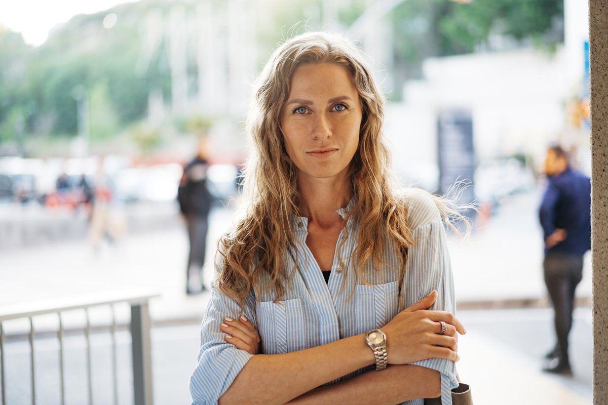 kobieta w wieku około 40 lat z długimi włosami blond falowanymi i w koszuli stoi na ulicy z założonymi rękami i poważną miną