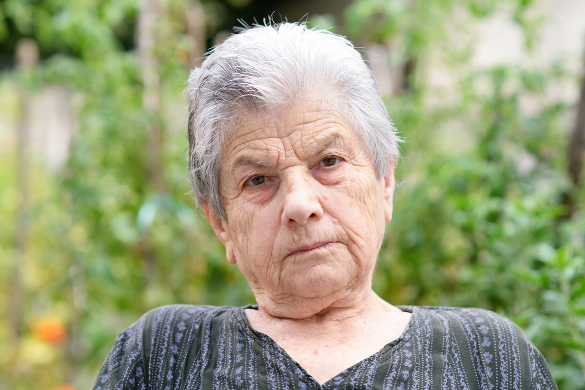 kobieta w senioralnym wieku 70+ z siwymi krótkimi włosami siedzi na zewnątrz na tle zieleni ze skwaszoną miną