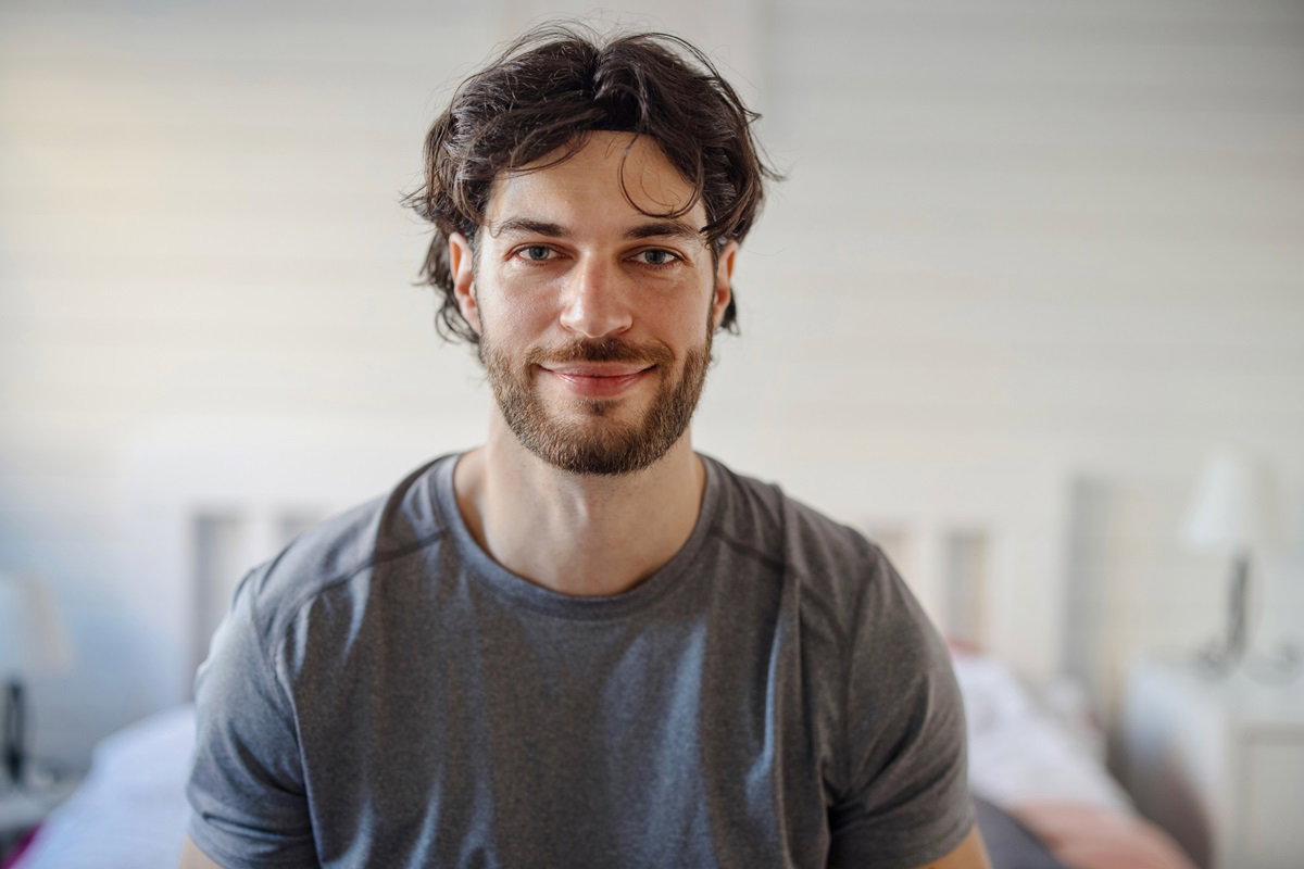 młody mężczyzna z ciemnymi lekko podrośniętymi włosami i z zarostem siedzi na łóżku i lekko się uśmiecha