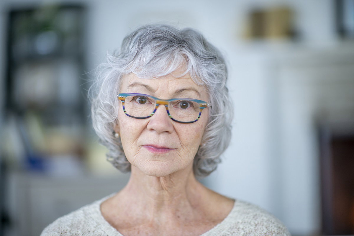 starsza kobieta z siwymi włosami i w okularach z kolorowymi oprawkami patrzy przed siebie smutnym wzrokiem