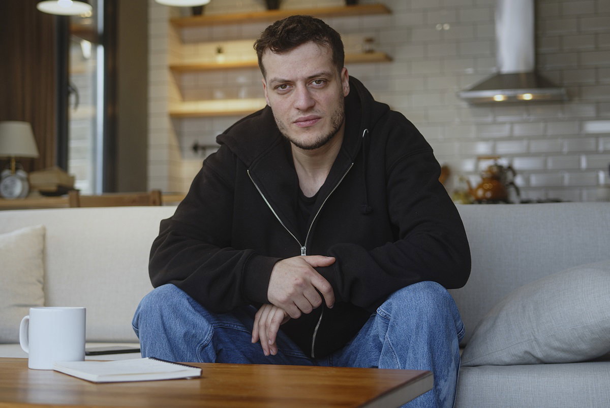 młody mężczyzna w wieku około 30 lat w czarnej bluzie i jeansach siedzi na kanapie na tle kuchni z wkurzoną miną