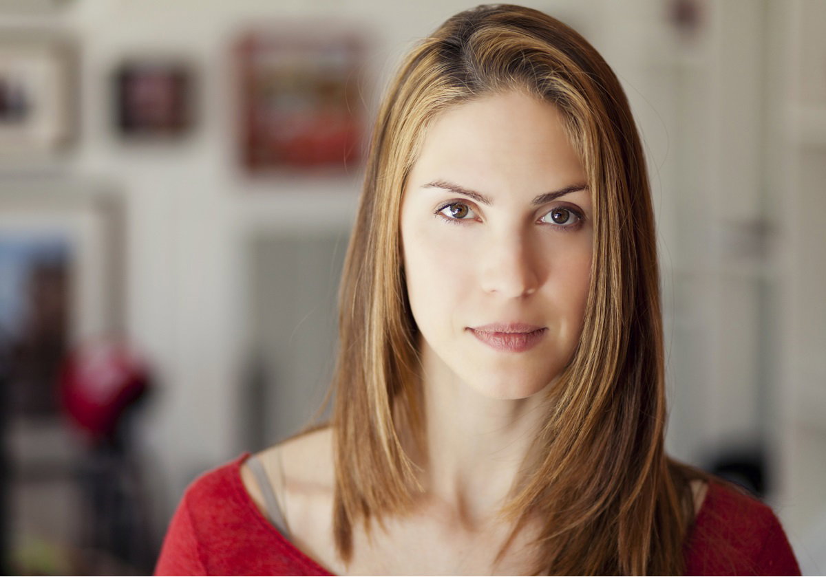 młoda kobieta w wieku około 30 lat w czerwonej bluzce stoi w salonie z poważną lekko pokpiwającą miną