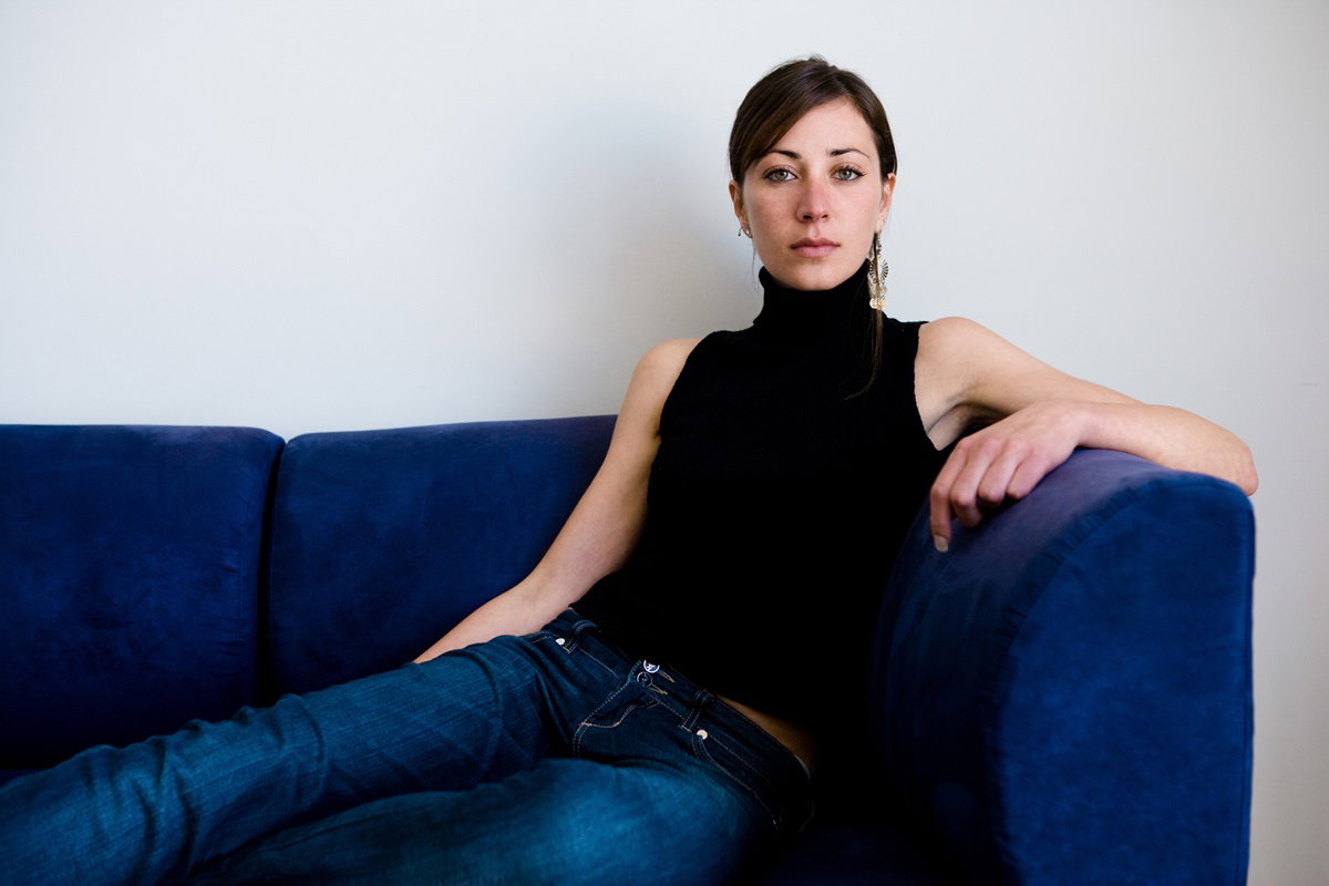 młoda kobieta w wieku około 35 lat z upiętymi włosami i w czarnym golfie bez rękawów i jeansach siedzi na kanapie z poważną miną