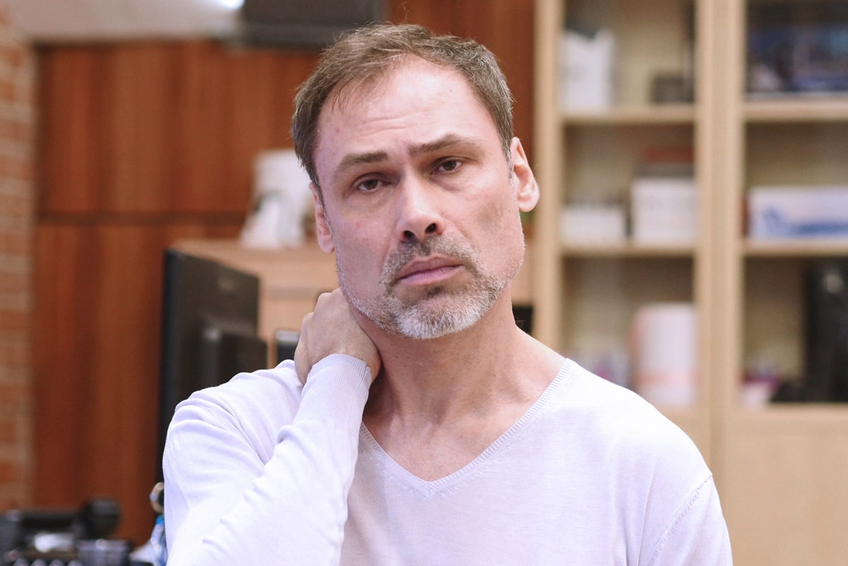 mężczyzna w wieku około 35 plus siwiejący i z zarostem w białej koszulce z długim rękawem stoi w salonie i jest zmartwiony