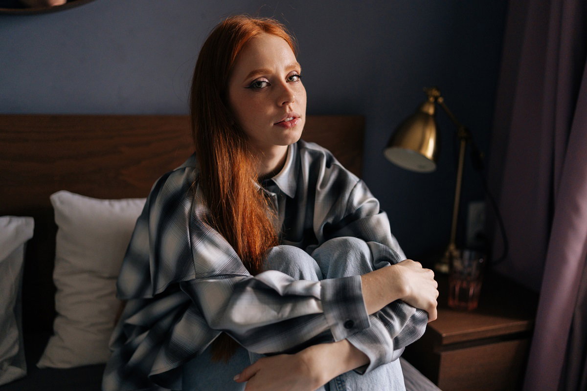 młoda dziewczyna w wieku około 28 lat w koszuli i z długimi rudymi włosami siedzi na łóżku i patrzy zasmucona