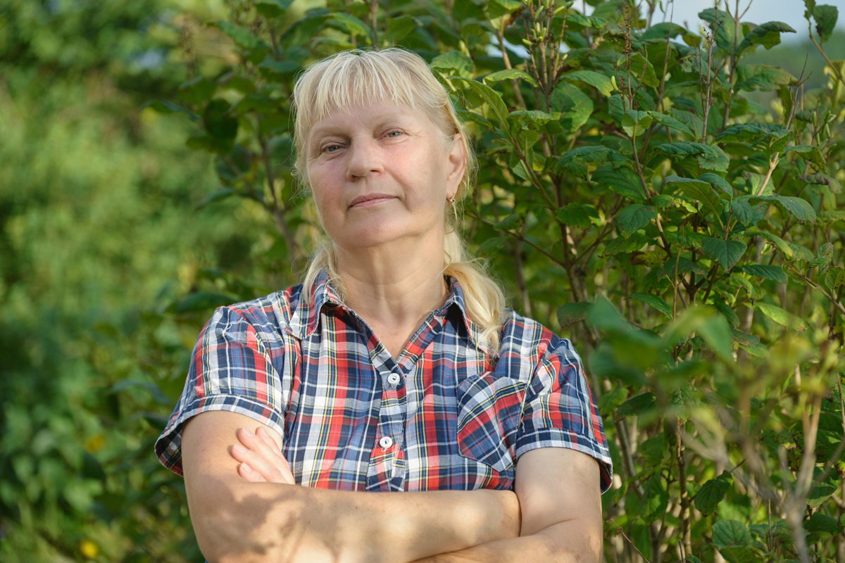 kobieta w wieku około 60 lat w koszuli w kratę blondynka stoi na tle zieleni z powątpiewającą miną