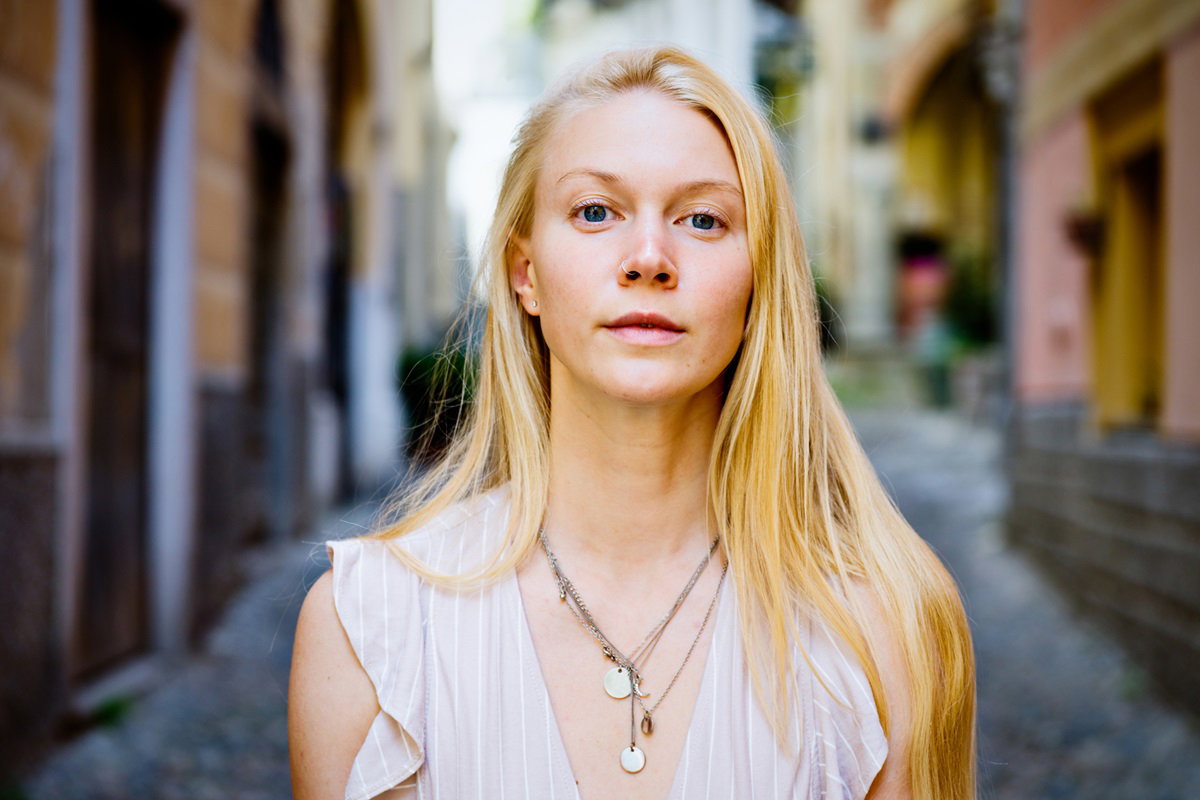 młoda dziewczyna w wieku około 30 lat z jasnymi długimi włosami i kolczykiem w nosie stoi na ulicy w letniej sukience i patrzy lekko wyzywająco
