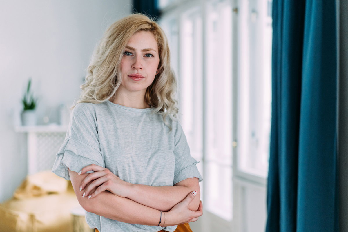 młoda kobieta z długimi blond włosami i w szarej bluzce stoi w salonie przy oknie z założonymi rękami i ma poważną minę