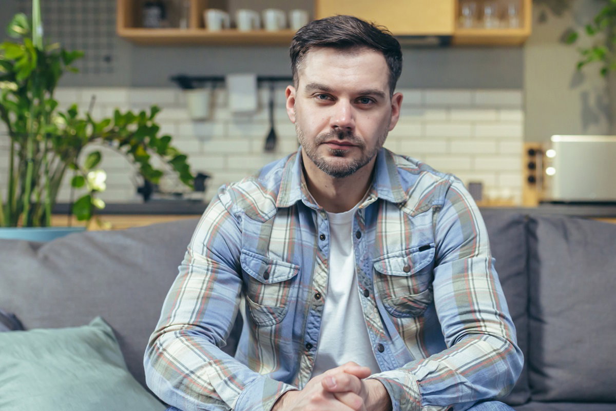 młody mężczyzna w wieku 30+ w koszuli w kratę siedzi na kanapie na tle kuchni i ma nietęgą minę