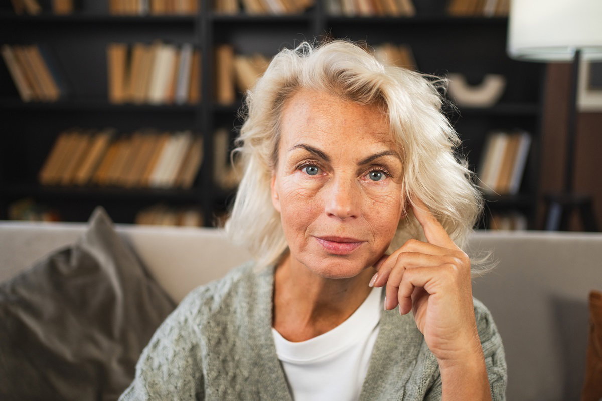kobieta w wieku 60 plus blondynka siedzi na kanapie w salonie i podpiera palcem skroń patrząc zamyślonym wzrokiem